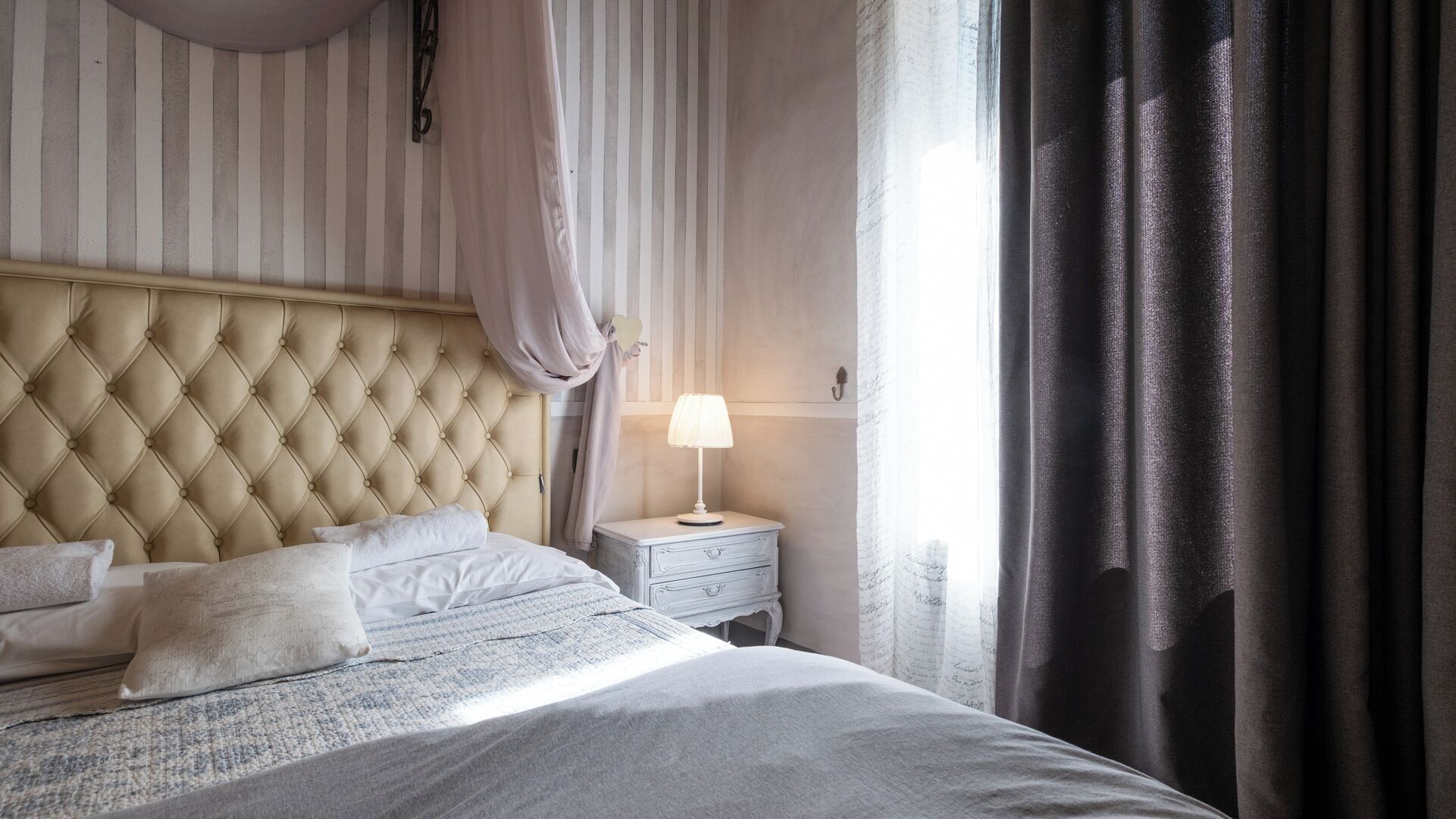 A neatly made bed with a tufted headboard, a bedside table with a lamp, and a window with curtains.