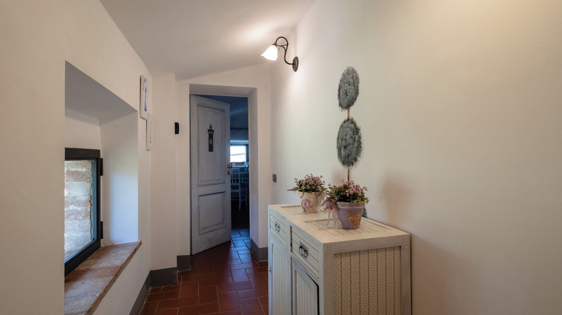 A hallway with a door leading to a room, a small window, and a cabinet with decorative items.