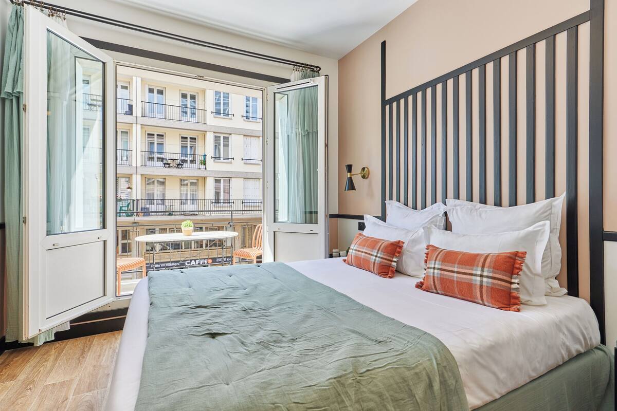 A hotel room with a large bed, a balcony with a table and chairs, and a view of other buildings.