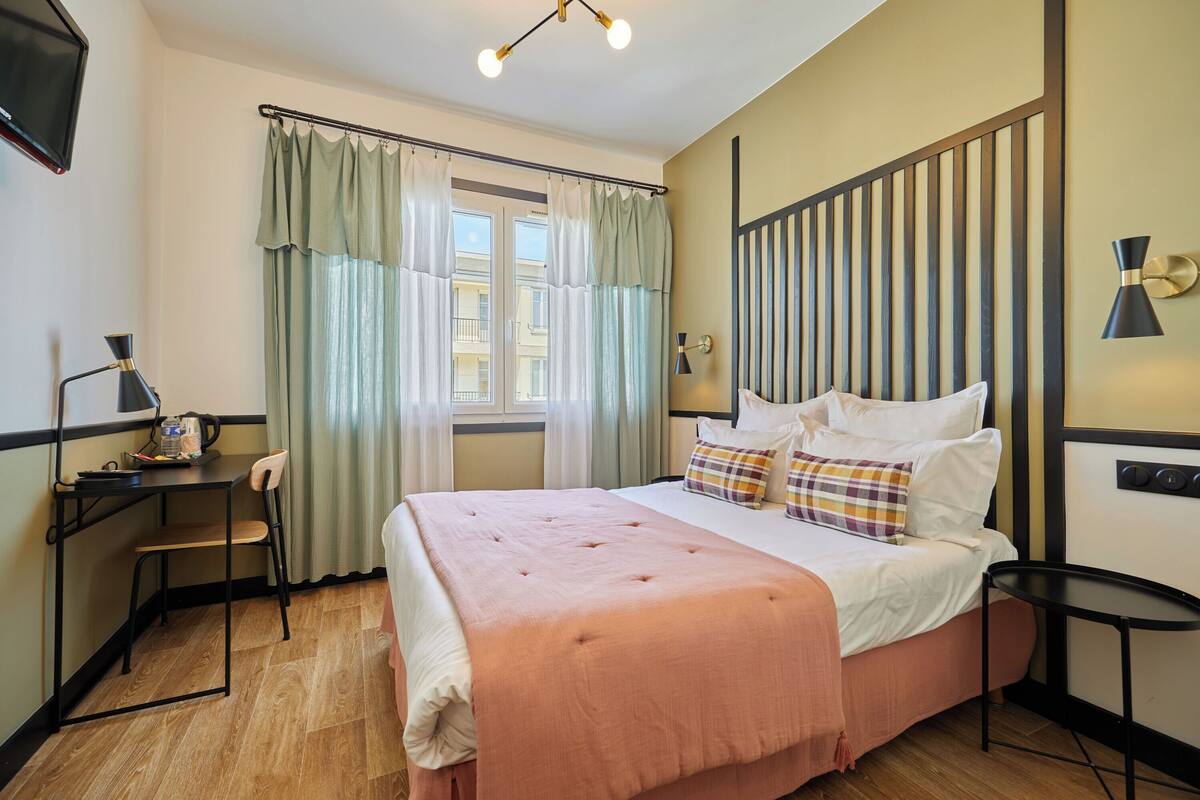 A hotel room with a bed, a desk, a chair, a TV, and a window with curtains.