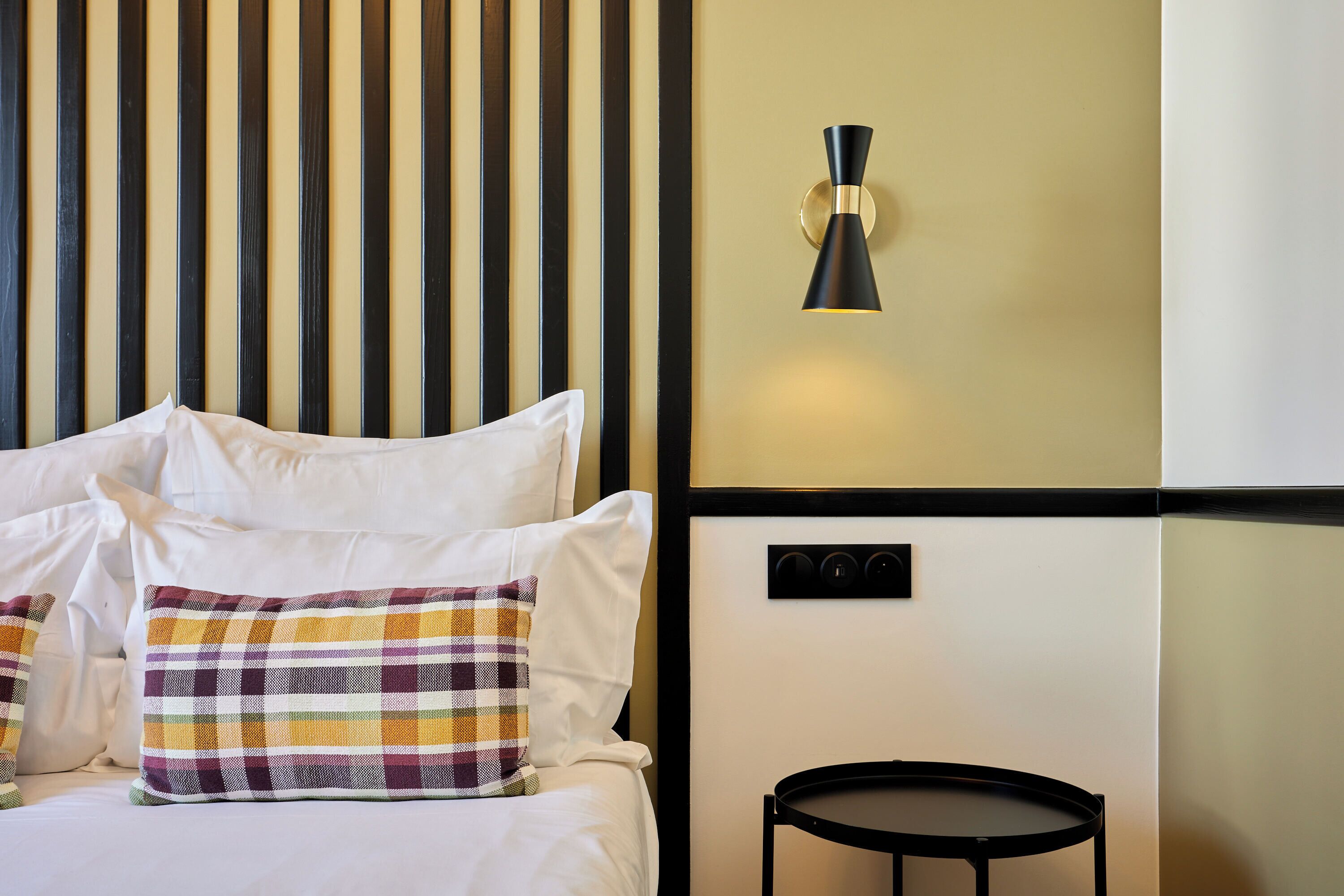 A neatly made bed with white linens and a plaid pillow, a black wall-mounted light fixture, and a small round table.
