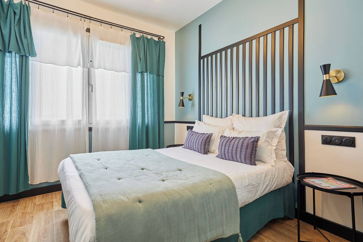 A neatly made bed with a white duvet and two pillows, a small bedside table with a magazine, and a wall-mounted lamp.