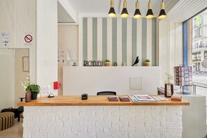 A reception desk with a white brick wall, a wooden counter, and a sign that reads "RICHELIEU". There are potted plants, a red fire alarm, and a framed picture on the counter. - Le Richelieu (Le Havre)