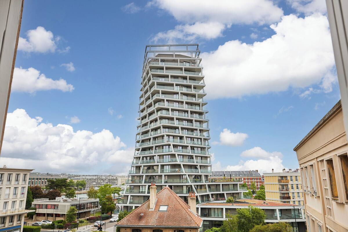 A modern high-rise building with multiple balconies, surrounded by other buildings and greenery.