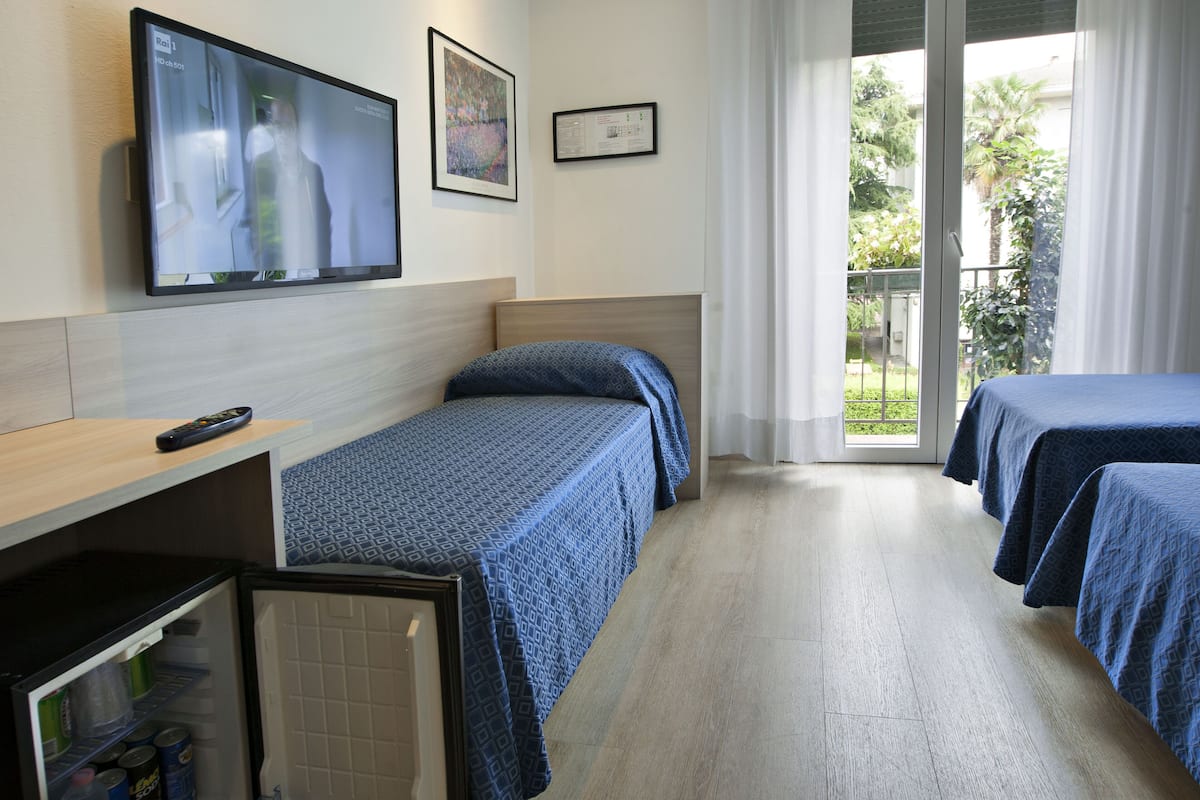 A hotel room with two beds, a TV mounted on the wall, a small refrigerator, and a window with a view of greenery.