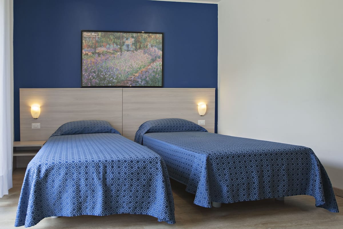 A room with two single beds, a wooden headboard, wall-mounted lights, and a framed painting above the bed.