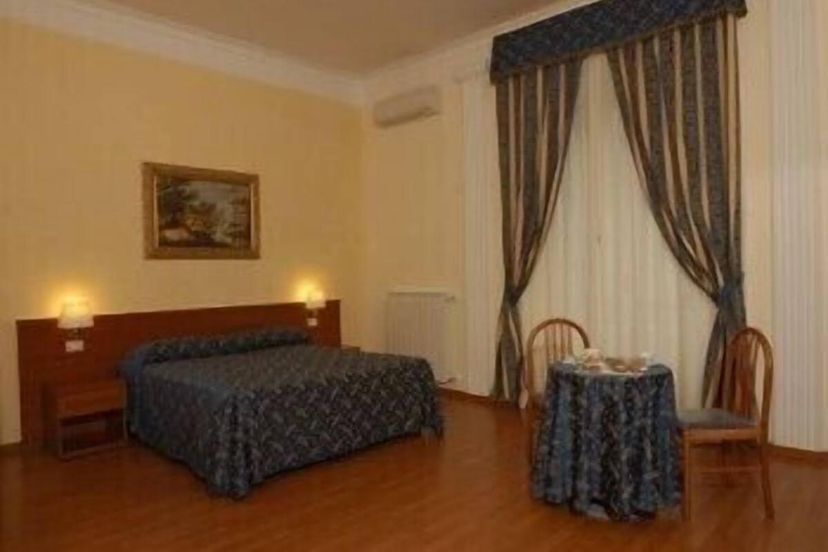 A bedroom with a bed, a table with chairs, a painting on the wall, and curtains.