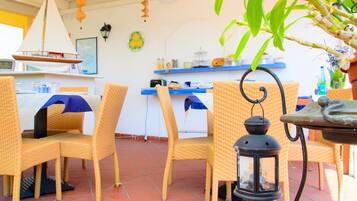 Uma sala de jantar com cadeiras de vime, uma mesa com um candeeiro e uma cozinha ao fundo.