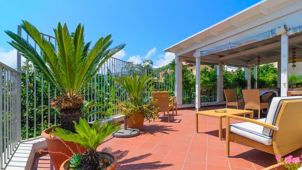 A covered outdoor seating area with wicker furniture, potted plants, and a view of the surrounding greenery. - Albergo Villa Giusto (Barano d'Ischia)