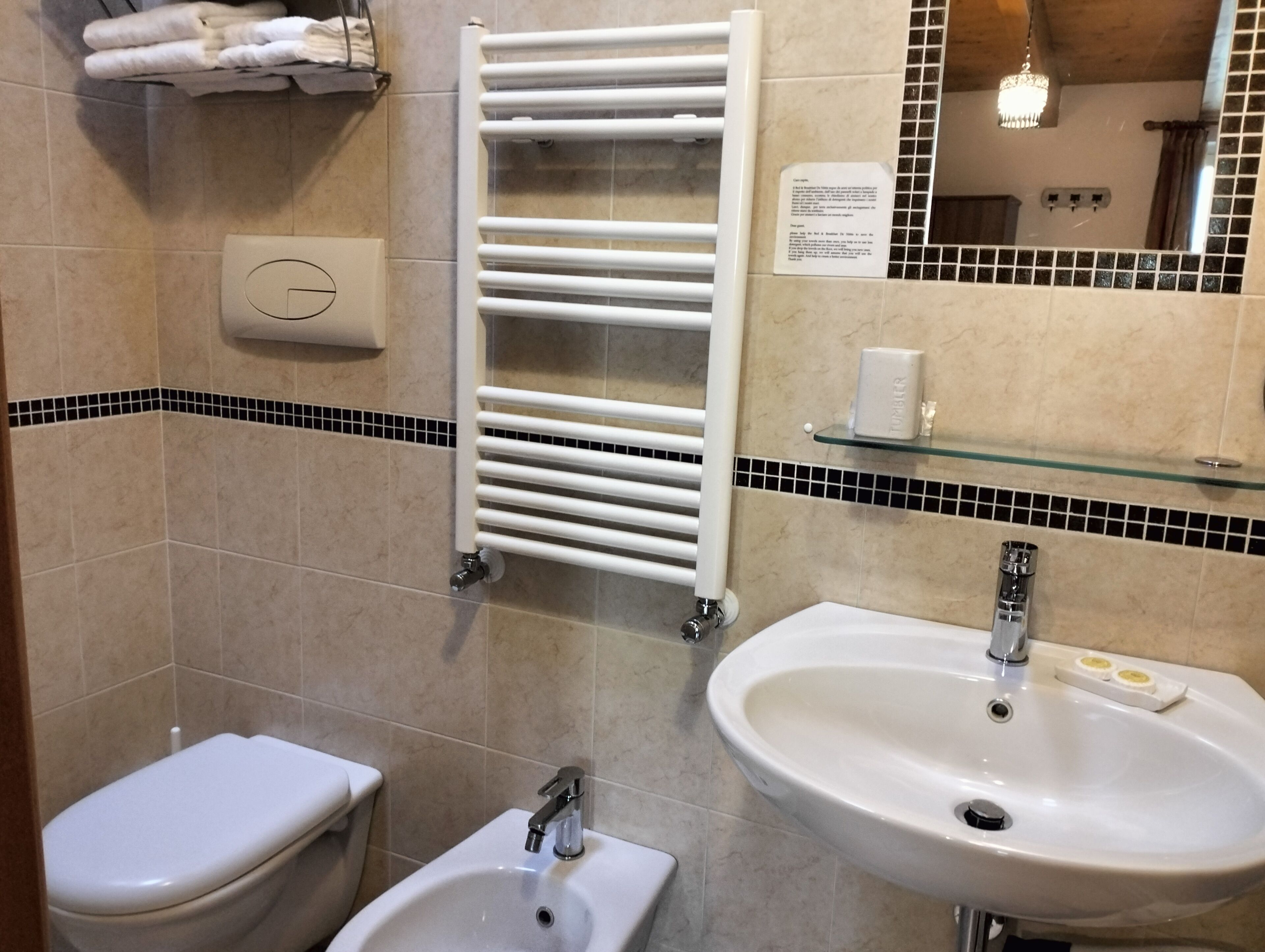 A bathroom with a toilet, bidet, and sink. There is a towel rack with folded towels, a glass shelf with toiletries, and a mirror with a chandelier.