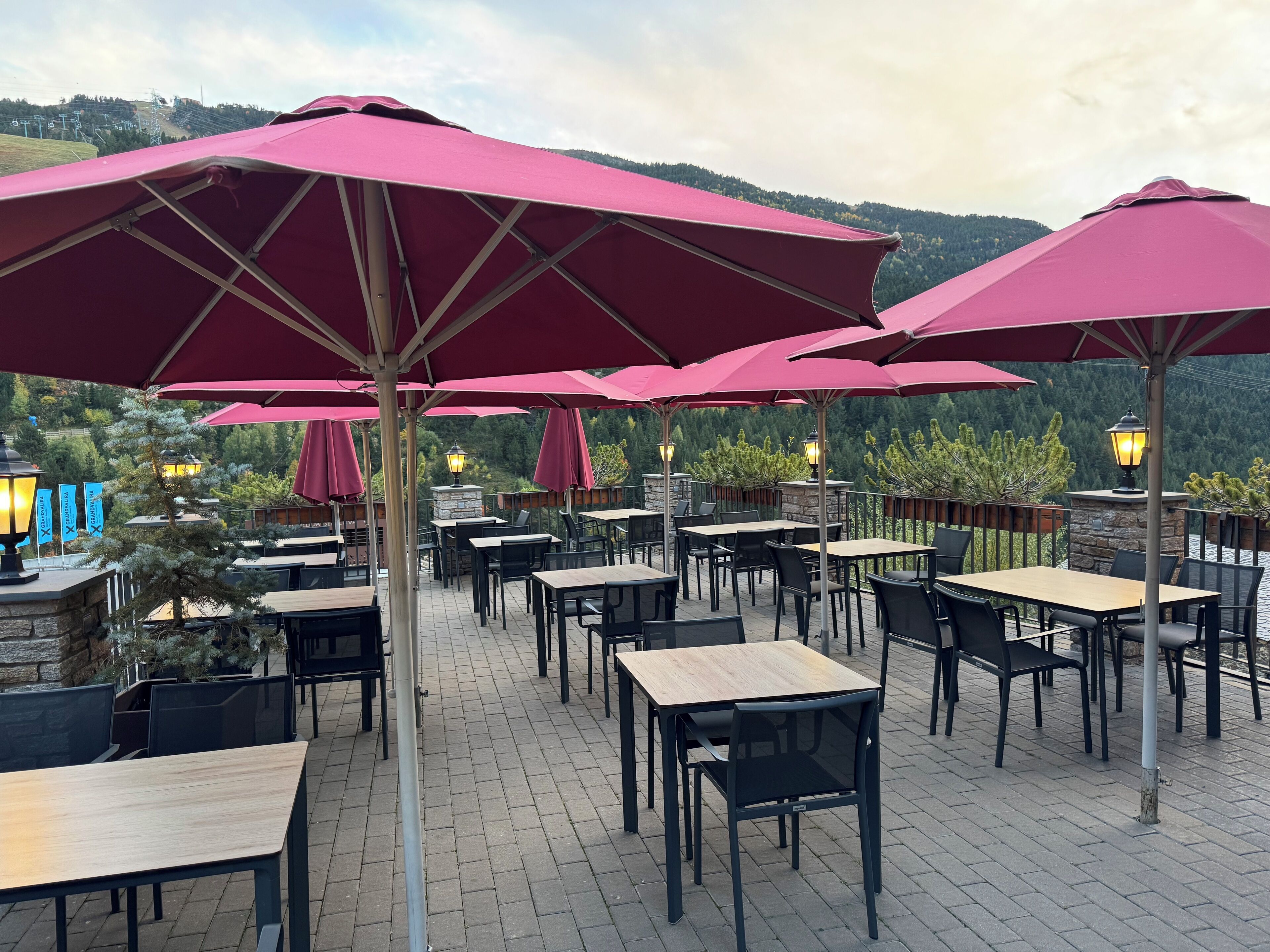 An outdoor patio area with tables and chairs, red umbrellas, and a view of a forested hillside.