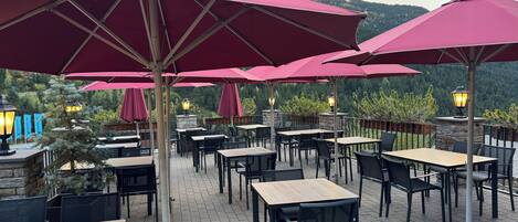 An outdoor patio area with tables and chairs, red umbrellas, and a view of a forested hillside.
