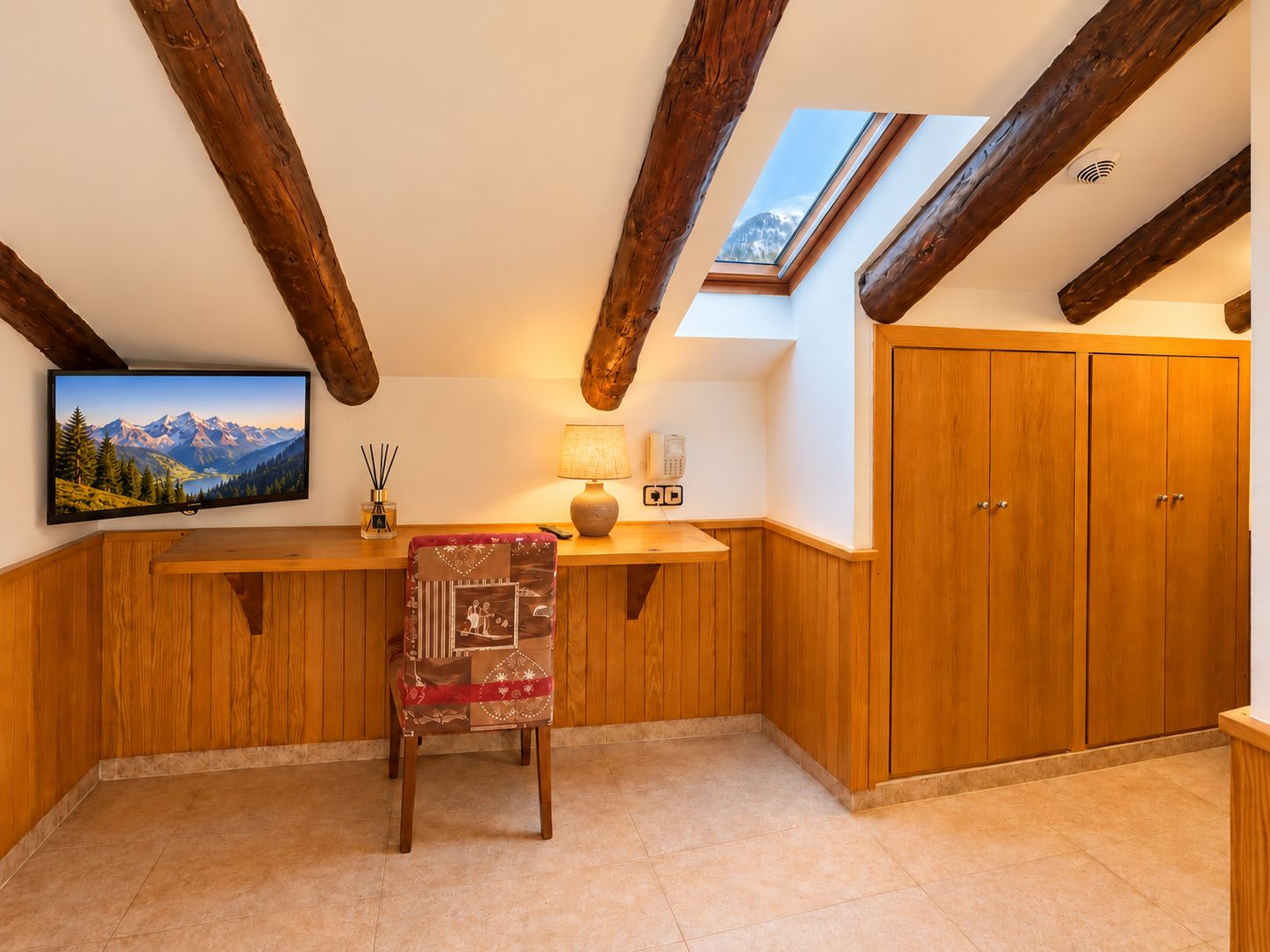 Una habitación con techo de madera, un televisor en la pared, un escritorio con lámpara, una silla y un armario.