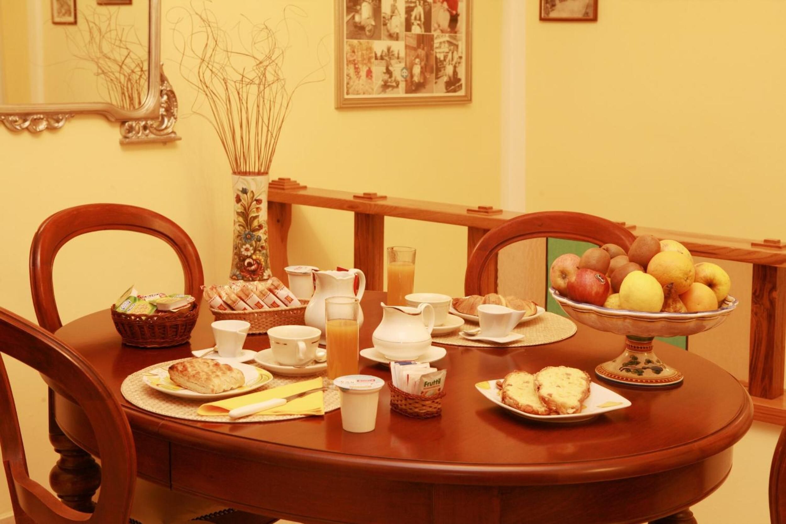 Una mesa de comedor lista con productos para el desayuno, como pan, bollería, fruta y bebidas.