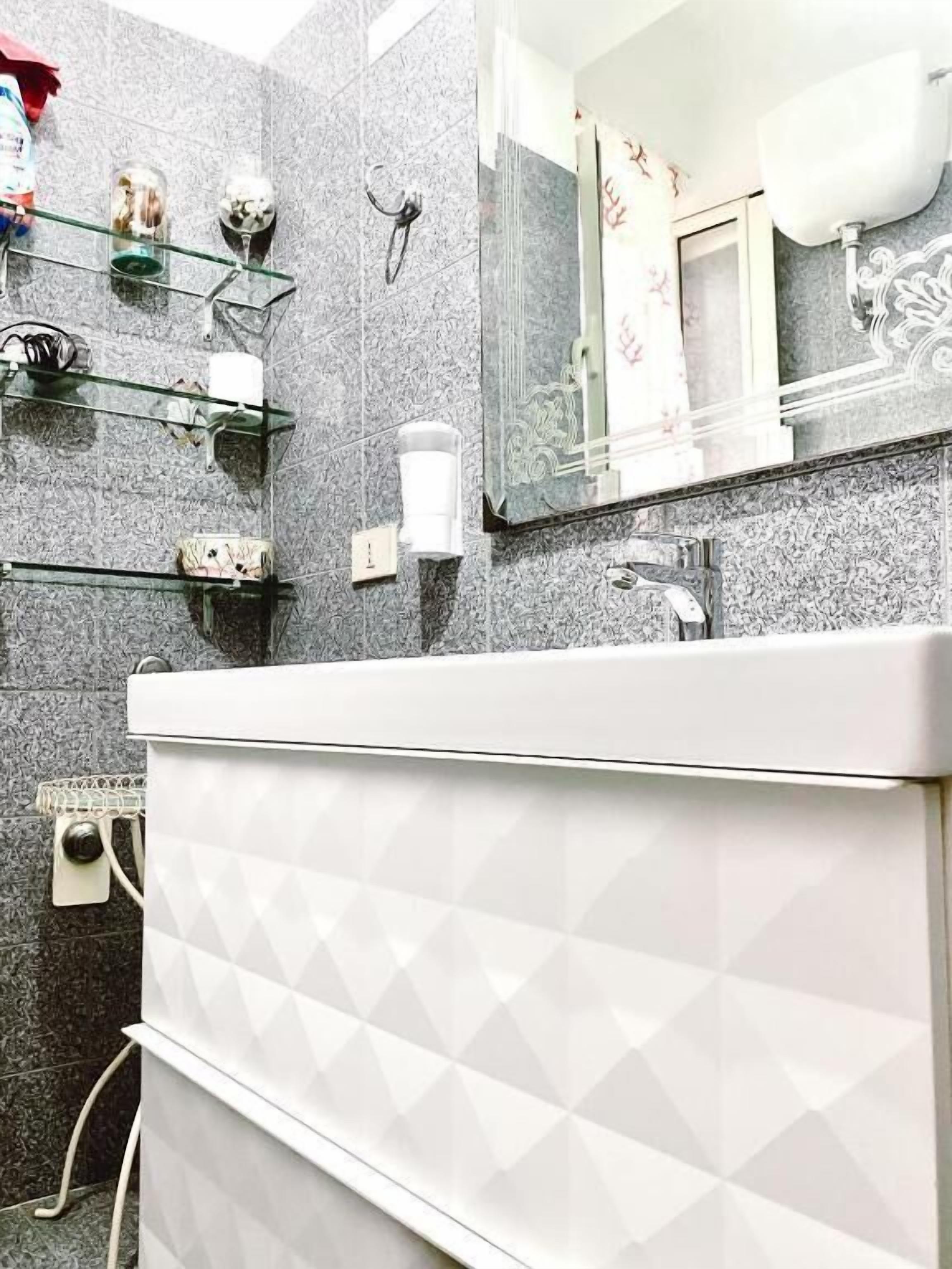 A bathroom with a white vanity, a glass shelf with toiletries, a mirror with decorative elements, and a textured wall.