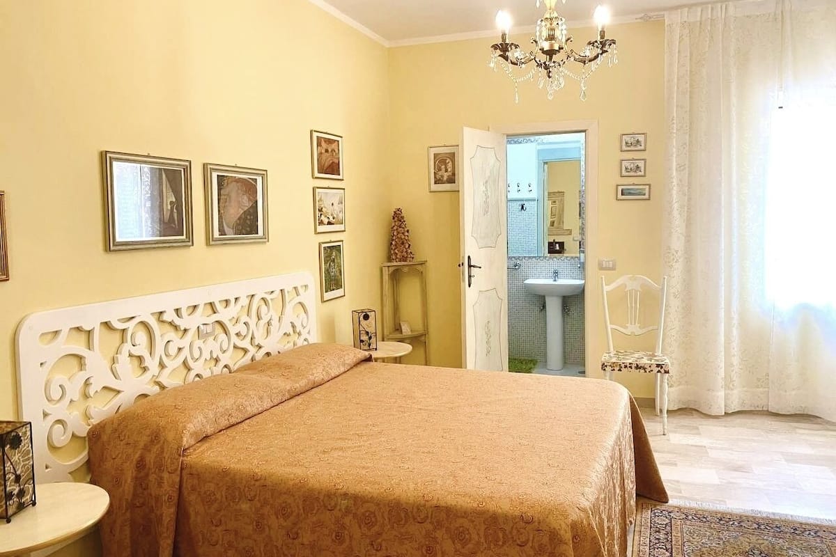 A bedroom with a large bed, a chandelier, a bathroom visible through an open door, and framed pictures on the wall.
