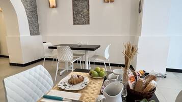 A dining area with a table set for breakfast, including croissants, fruits, and coffee. There are white chairs and a modern wall with artwork.