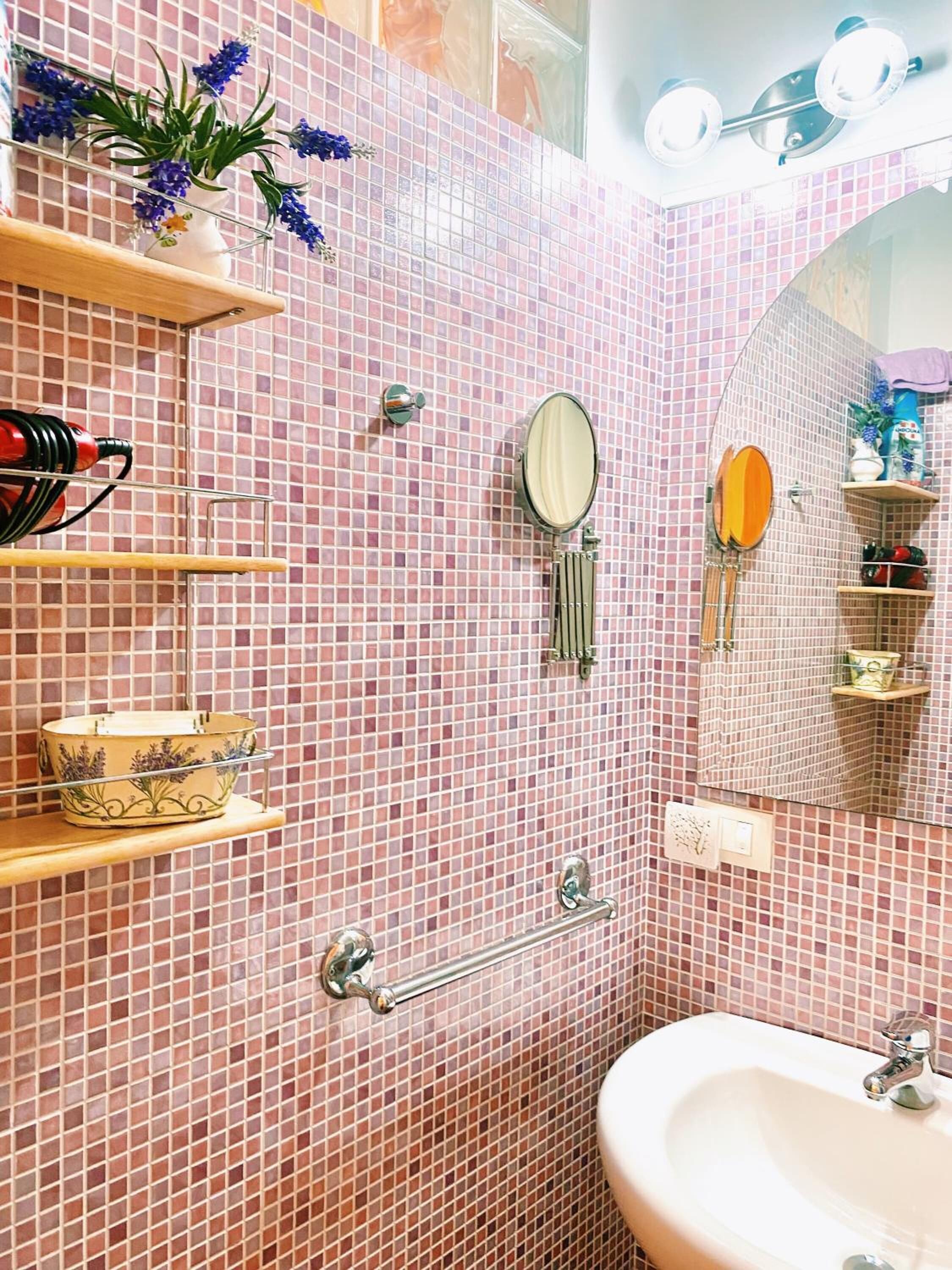 Un baño con mosaicos rosas, un lavabo, un espejo y repisas con artículos de aseo.