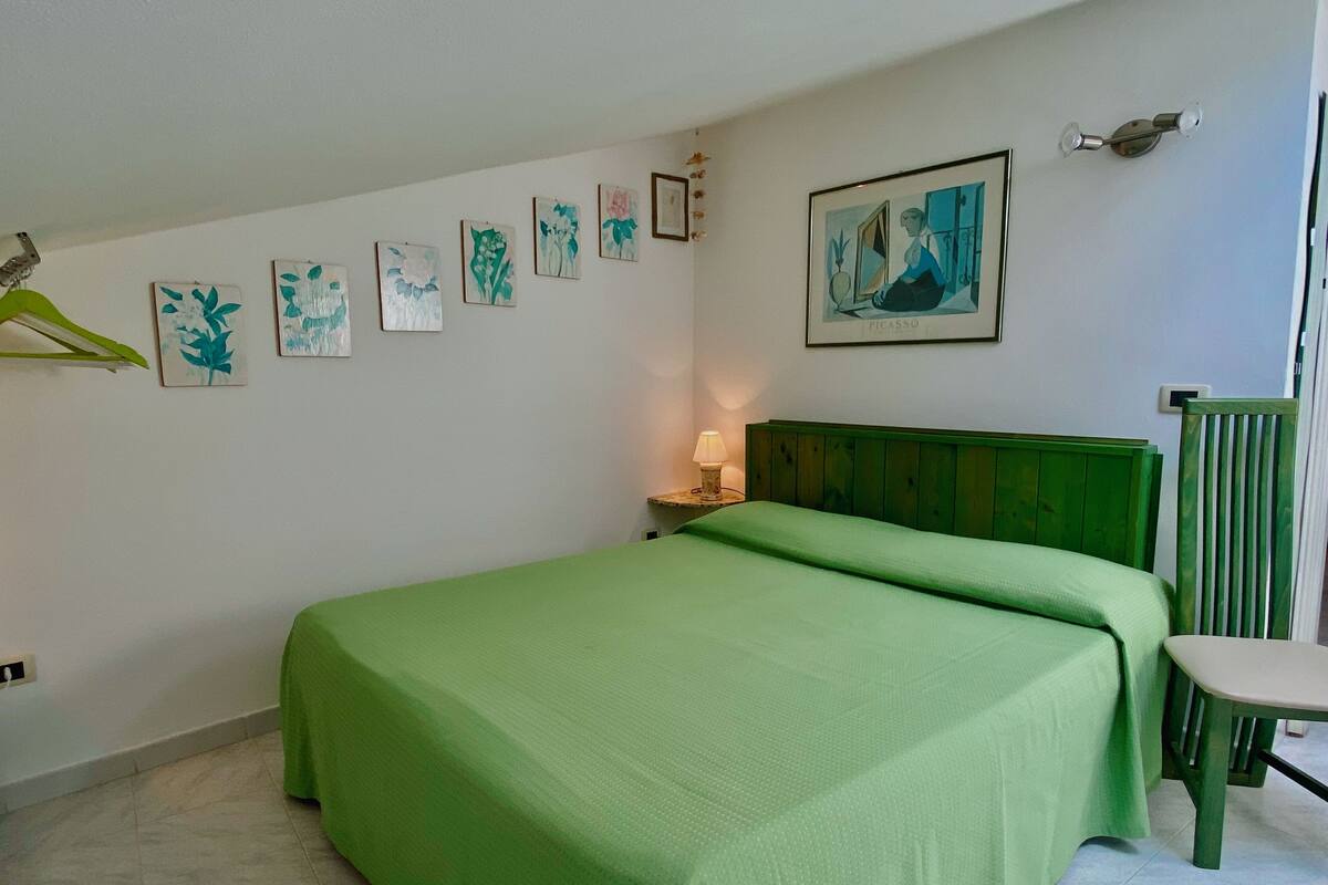 A bedroom with a green bedspread, a nightstand with a lamp, a chair, and several framed artworks on the wall.