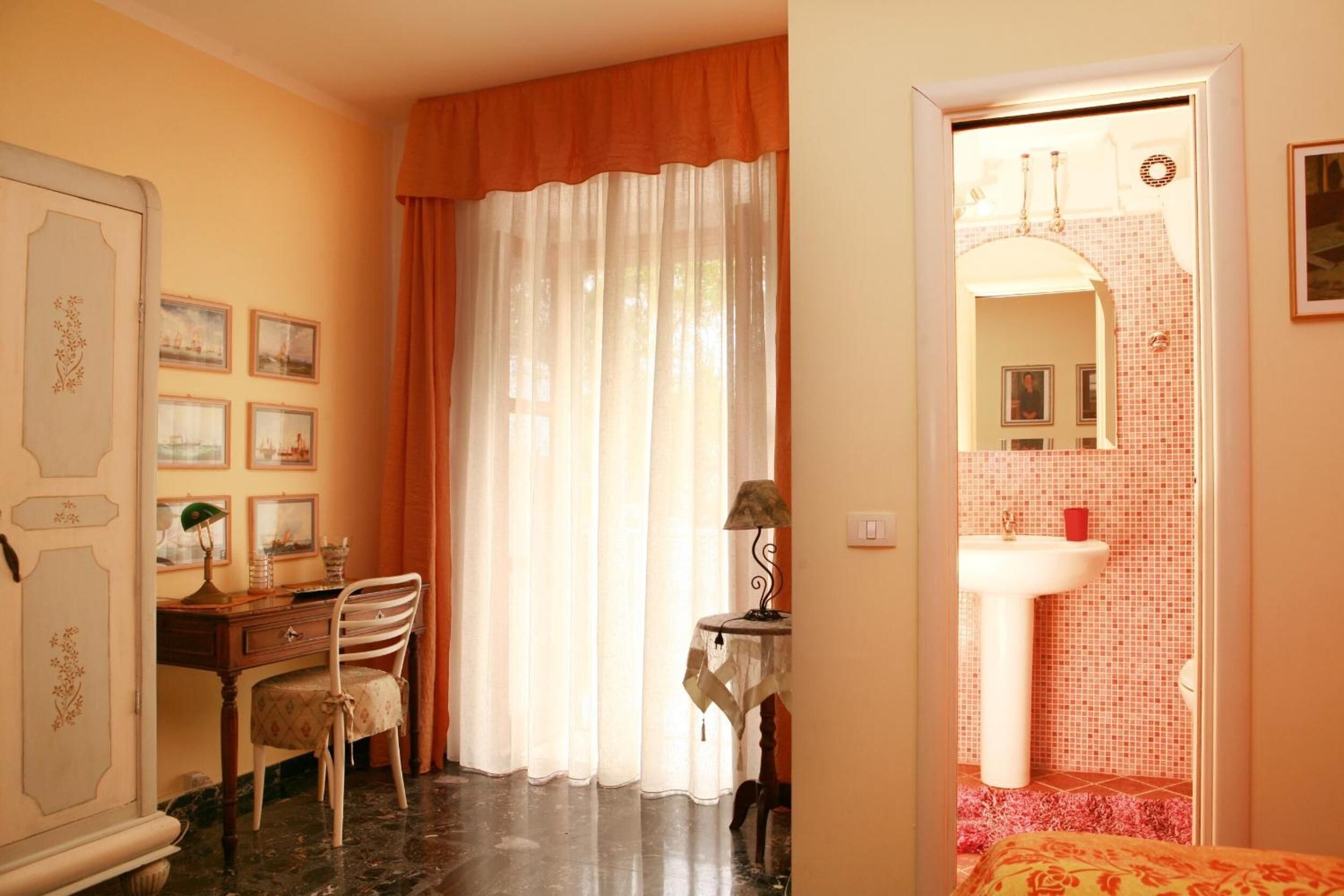 Ein Zimmer mit Schreibtisch, Stuhl und Bett. Ein Fenster mit Vorhängen, eine Tür zu einem Badezimmer und eine Wand mit gerahmten Bildern.