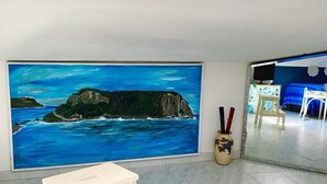 A room with a large painting of a coastal landscape, a white table, and a vase with red and black objects. - Al Vecchio Pioppo (Praia a Mare)
