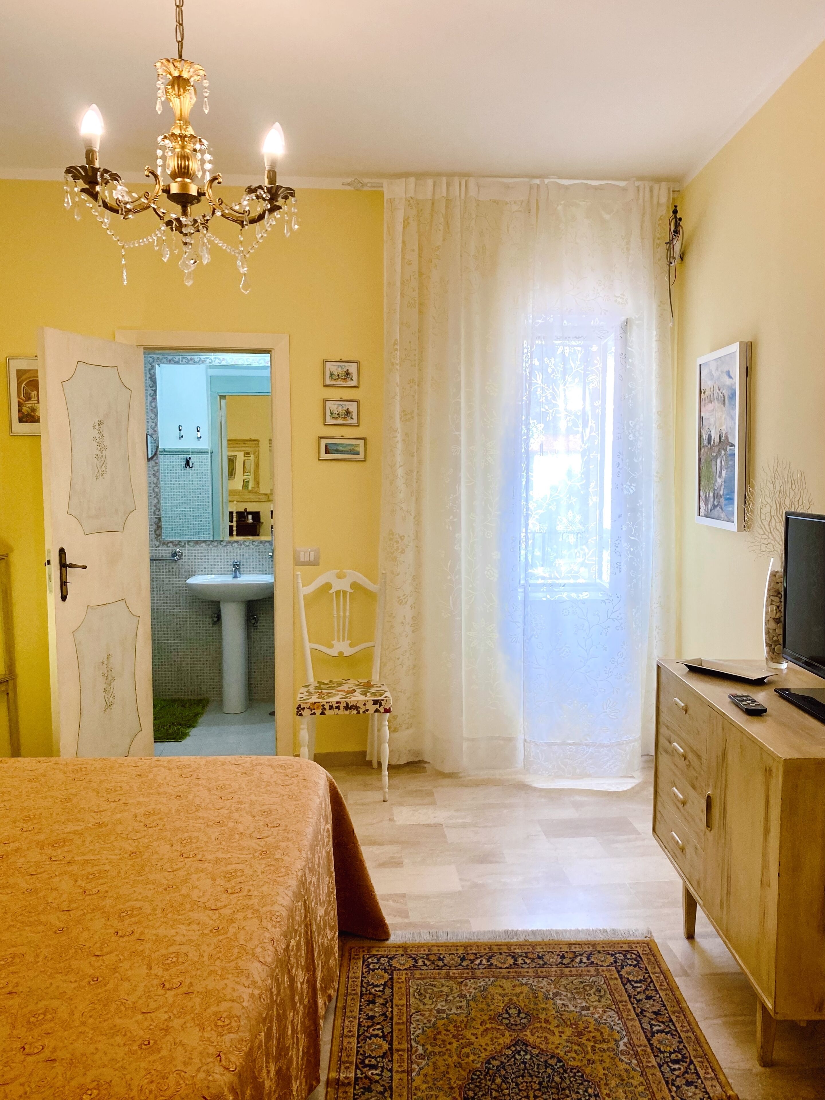 Una camera da letto con un letto, una cassettiera in legno, una televisione e un bagno visibile attraverso una porta aperta.