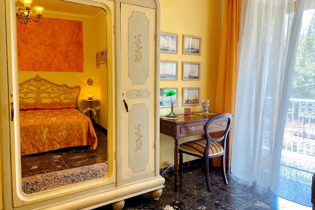 A bedroom with a bed, a desk, and a chair. There are framed pictures on the wall and a window with curtains.