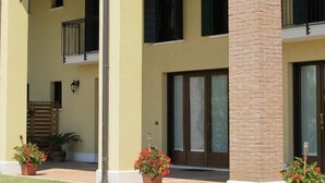 A multi-story building with a yellow facade, black shutters, and a brick column. There are potted plants in front of the entrance and a small balcony with a metal railing. - Ca' Florian (Zero Branco)