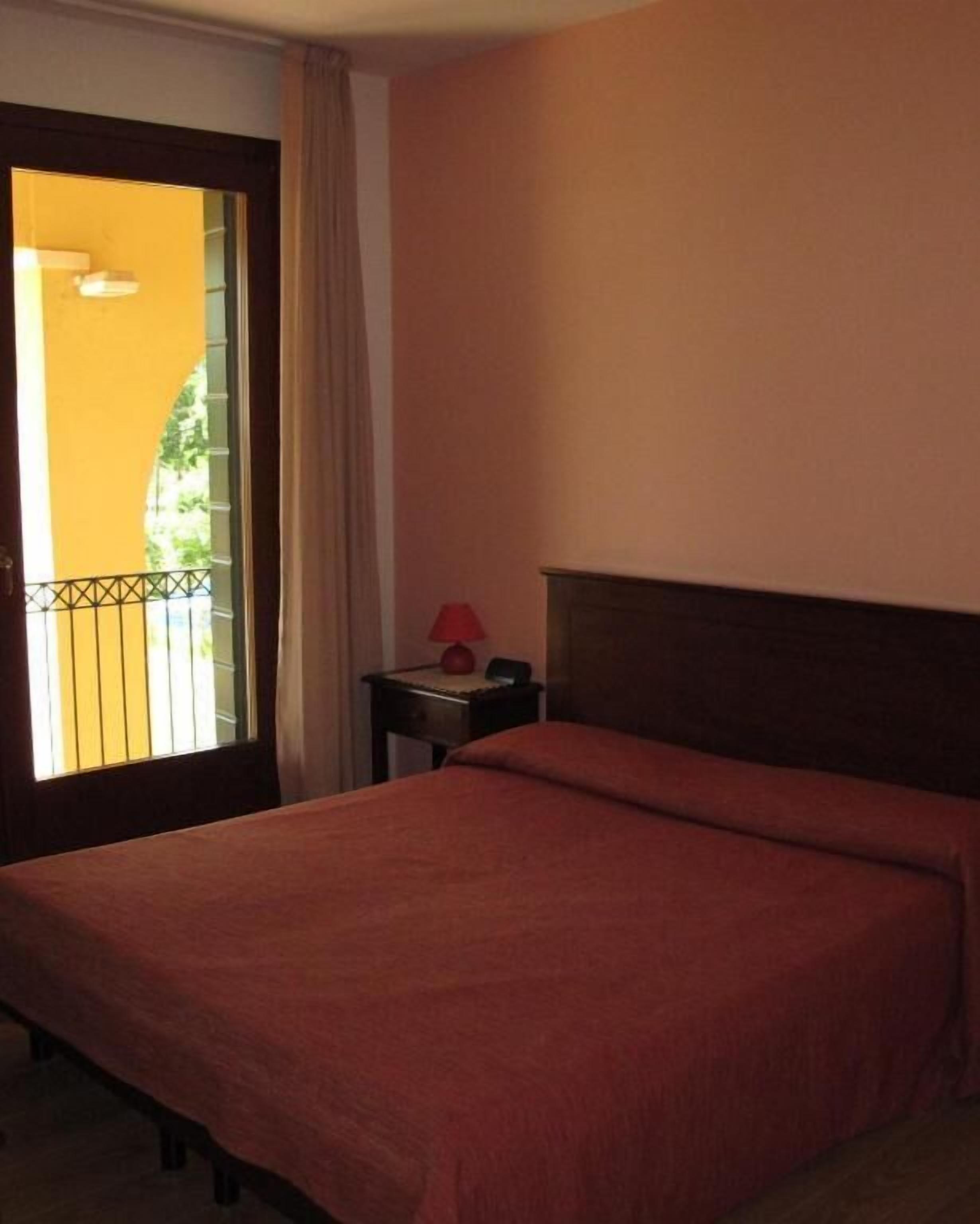 Habitación de hotel con cama, mesita de noche, lámpara y vistas al exterior.