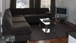 A modern living room with a sectional sofa, a small table, a television, and a view of a cityscape through large windows. - Dreamhouse Apartments Glasgow City Centre (Glasgow)