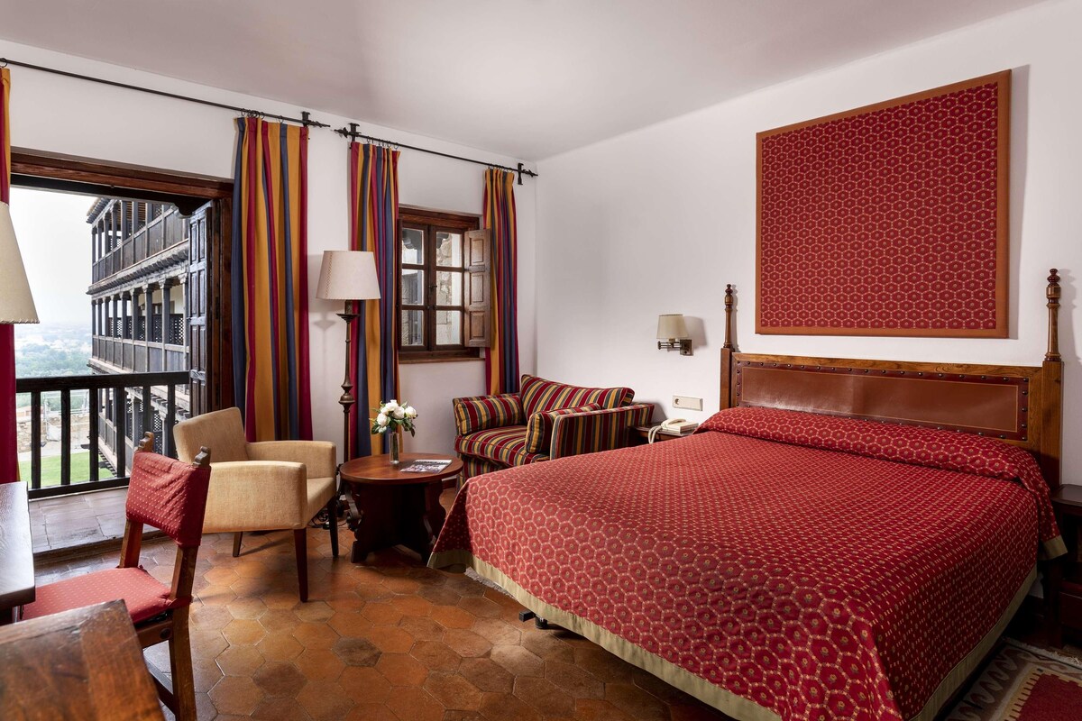Habitación de hotel con cama, una silla, una mesita, una lámpara y vistas al exterior.