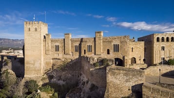 A historic stone castle with multiple towers and arched windows, surrounded by rocky terrain and sparse vegetation.