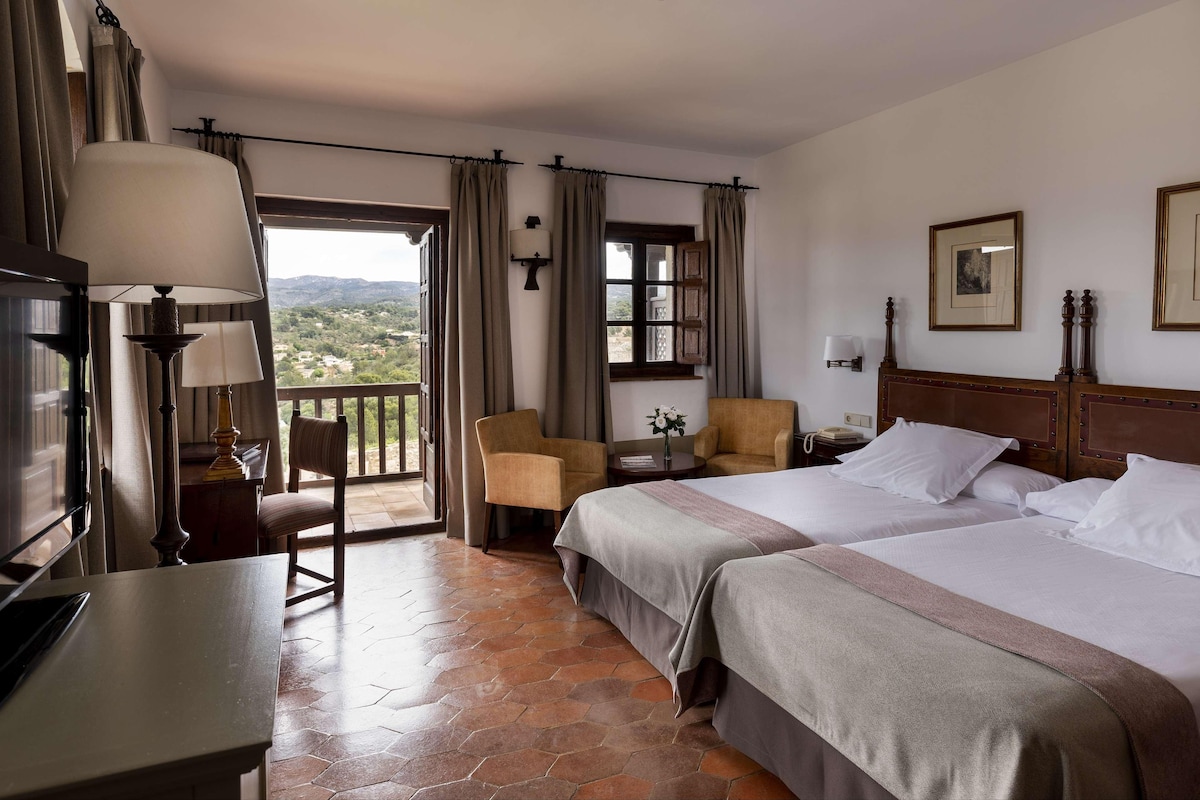 A hotel room with two beds, a desk, a chair, a lamp, and a view of the outdoors.