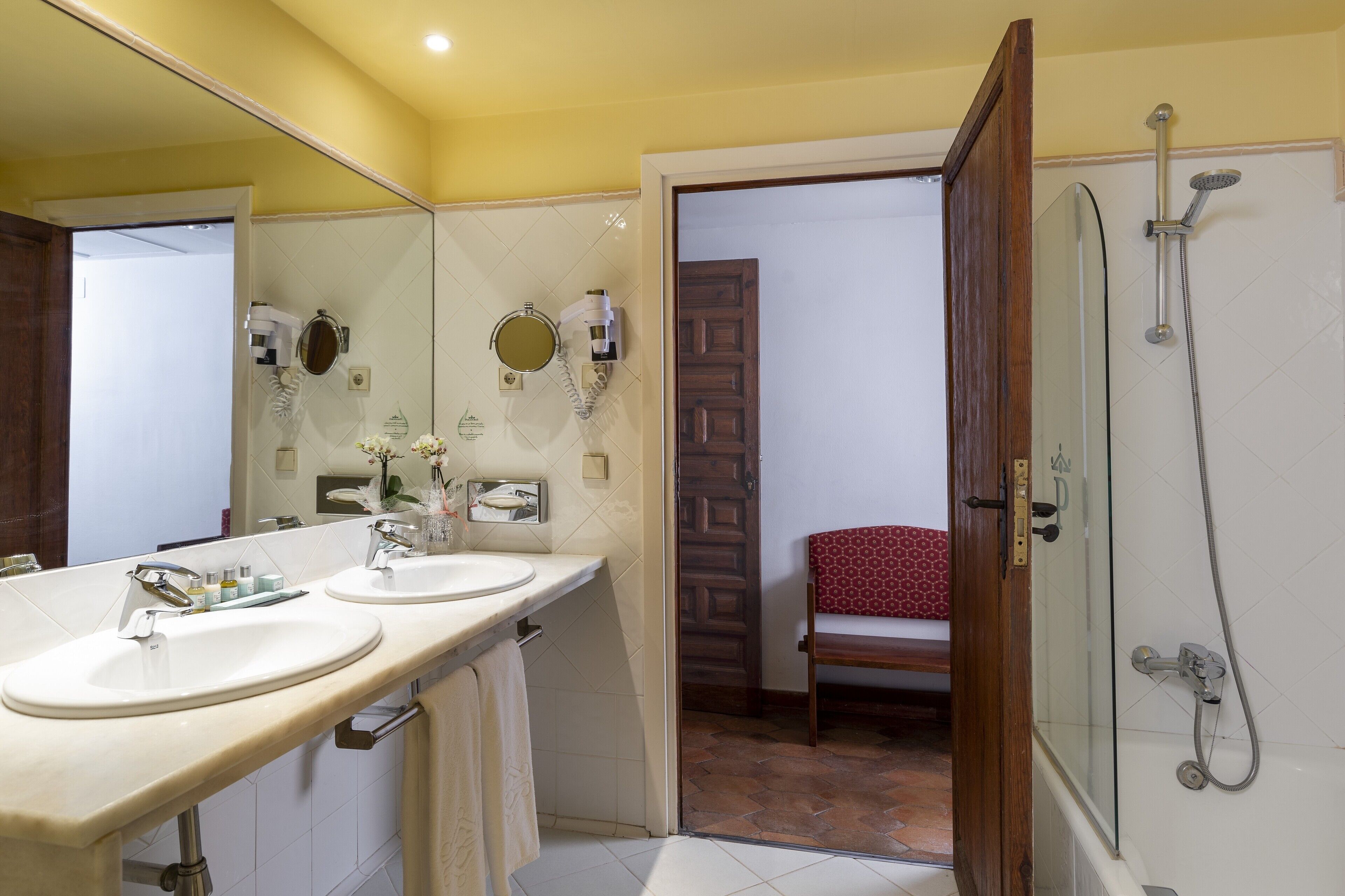Une salle de bain avec deux lavabos, un miroir, une douche et une porte donnant sur une chambre.