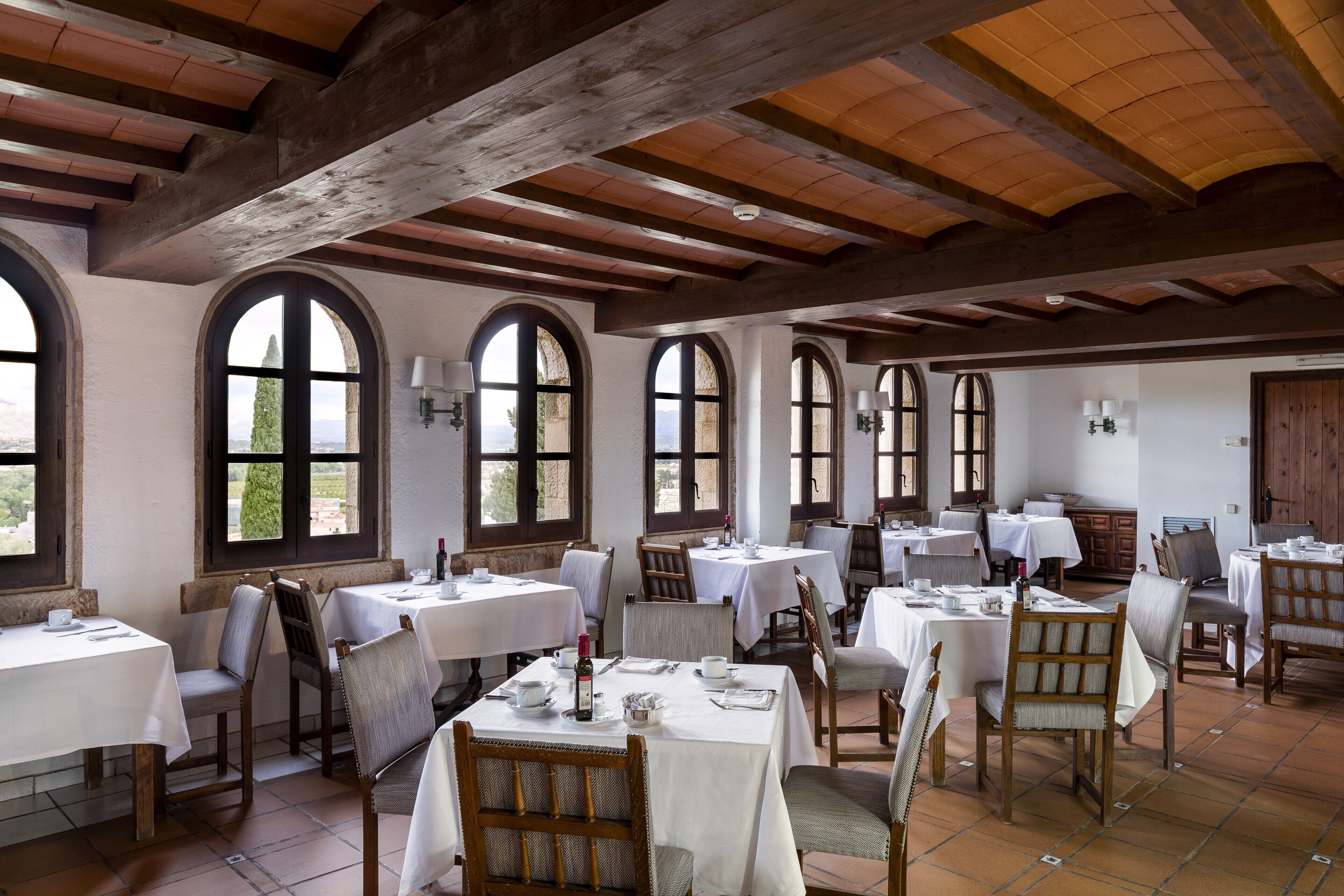 A dining area with multiple tables and chairs, large arched windows, and wooden beams on the ceiling.