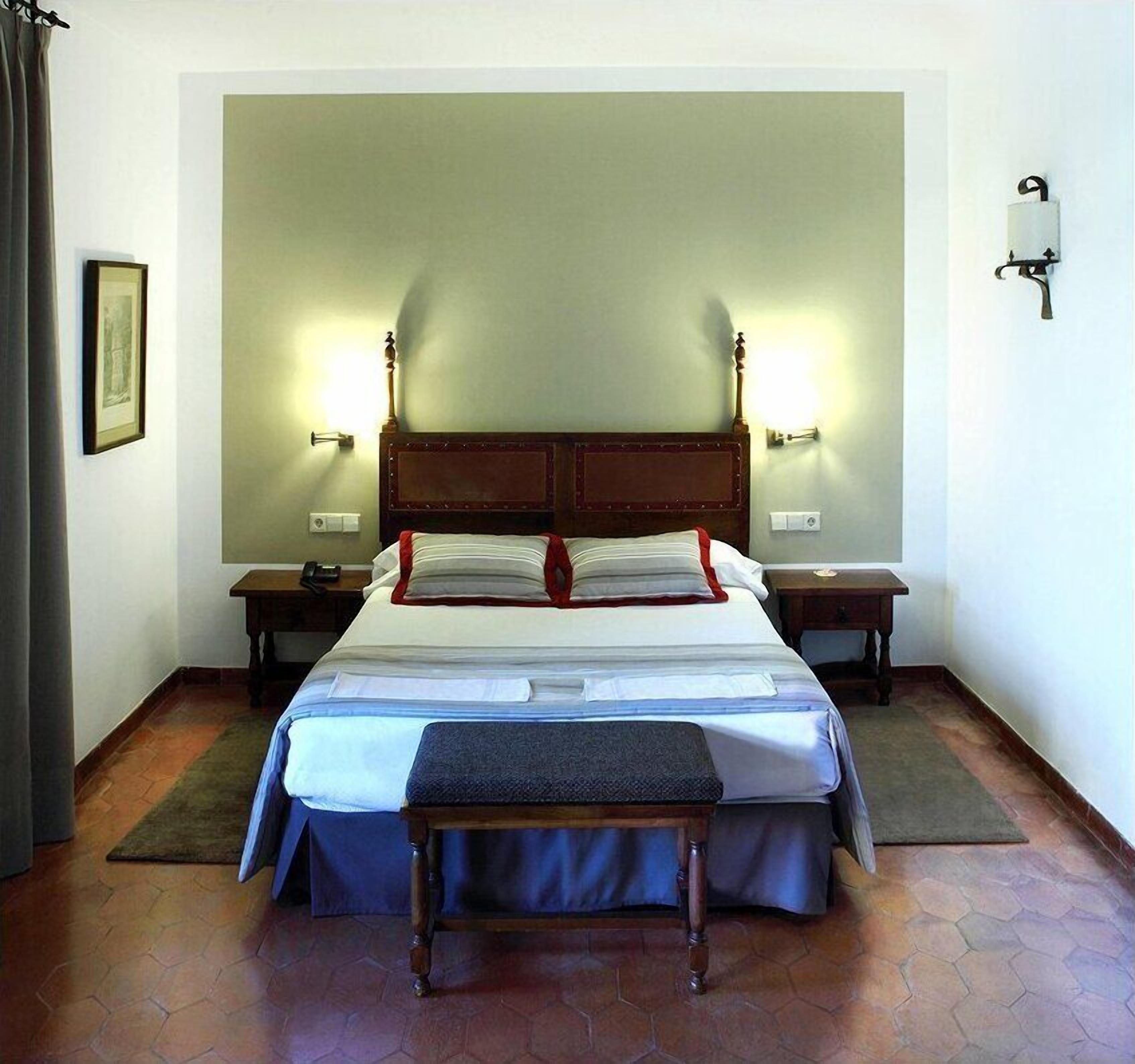 Une chambre d’hôtel avec un grand lit, deux tables de chevet, une petite table, un banc et des lampes fixées au mur.