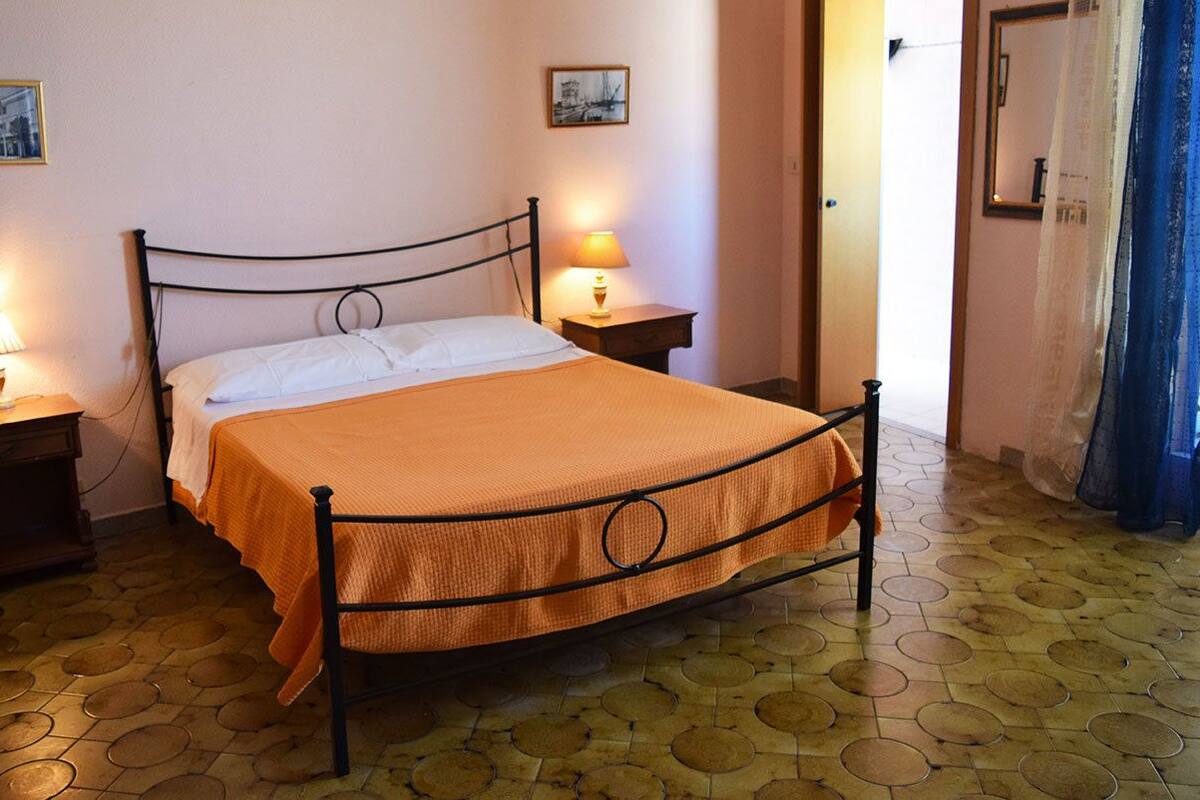 A single bed with an orange bedspread in a room with a tiled floor and two bedside tables with lamps.