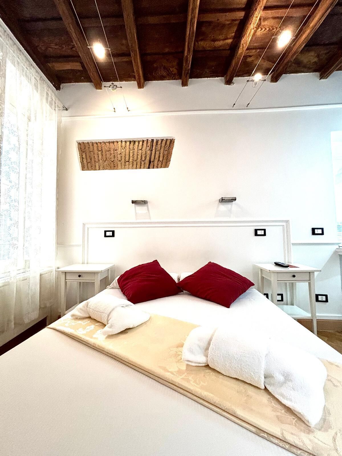 A bedroom with a large bed, two bedside tables, a wooden ceiling, and a window with curtains.