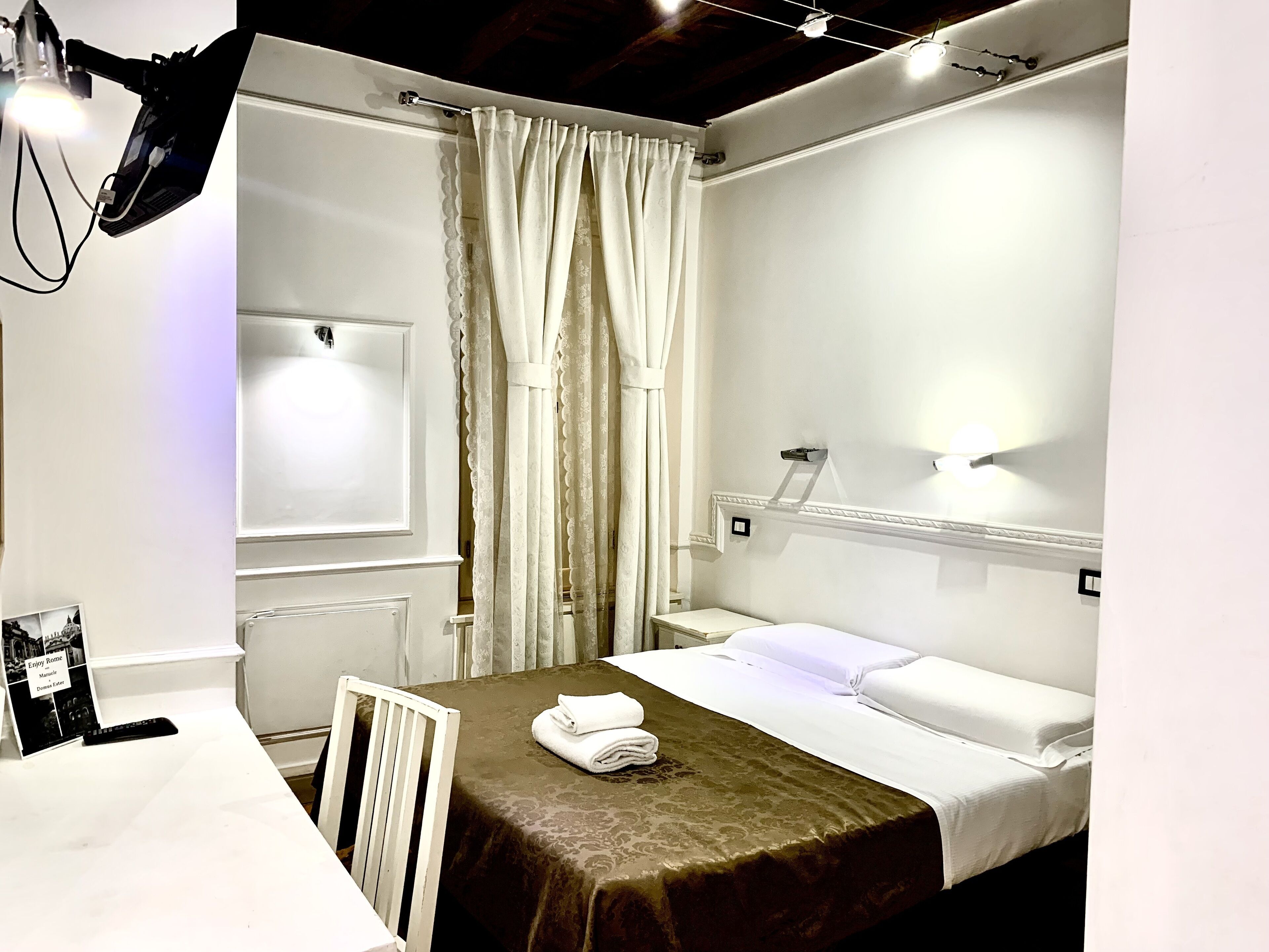 Une chambre d’hôtel avec un lit, un bureau et une chaise. Il y a une fenêtre avec des rideaux, un luminaire fixé au mur et un cadre photo accroché au mur.