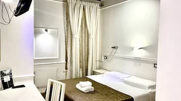 Une chambre d’hôtel avec un lit, un bureau et une chaise. Il y a une fenêtre avec des rideaux, un luminaire fixé au mur et un cadre photo accroché au mur.