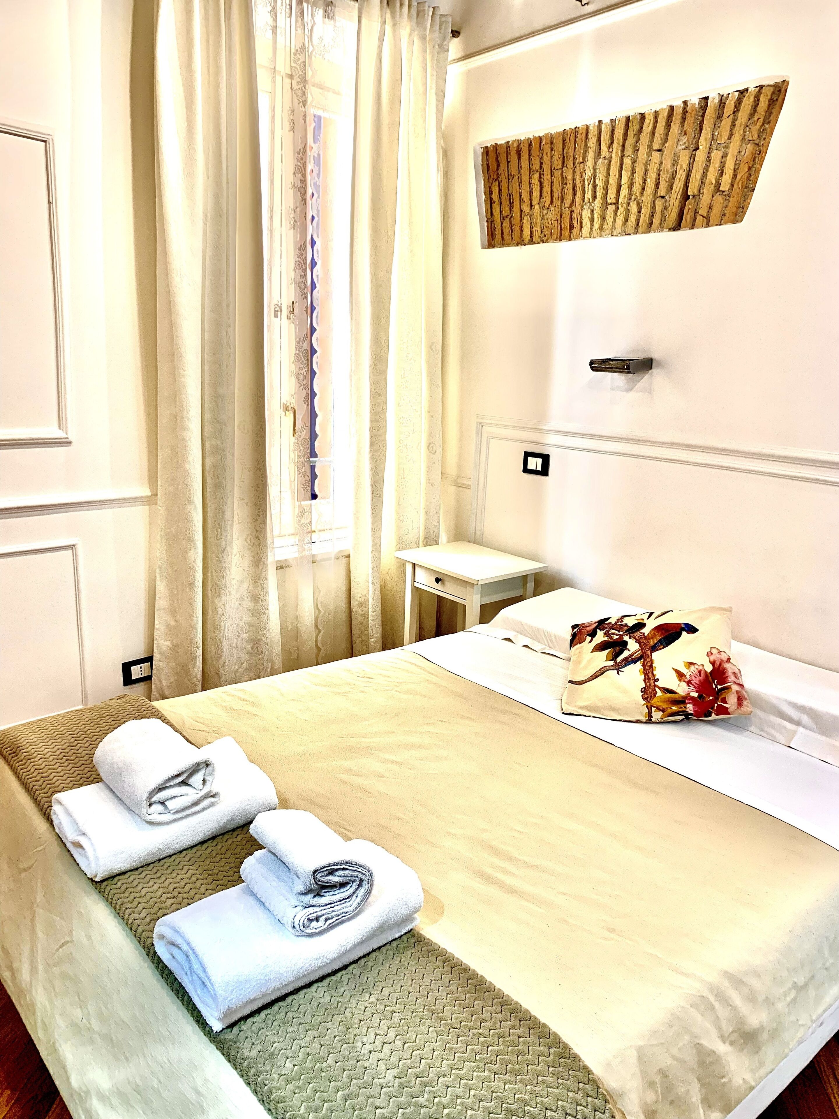 A hotel room with a bed, a small bedside table, a decorative pillow, and neatly folded towels.