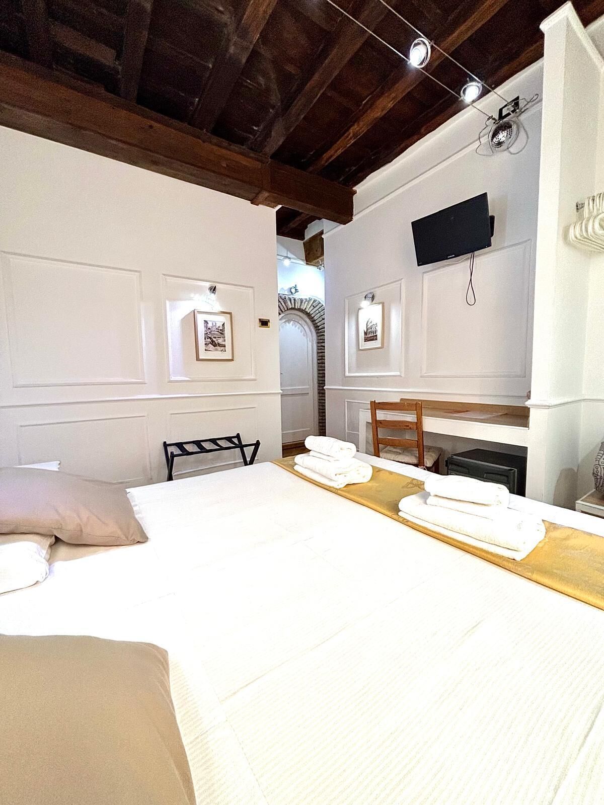 A hotel room with a bed, a wooden table with towels, a chair, and framed pictures on the wall.