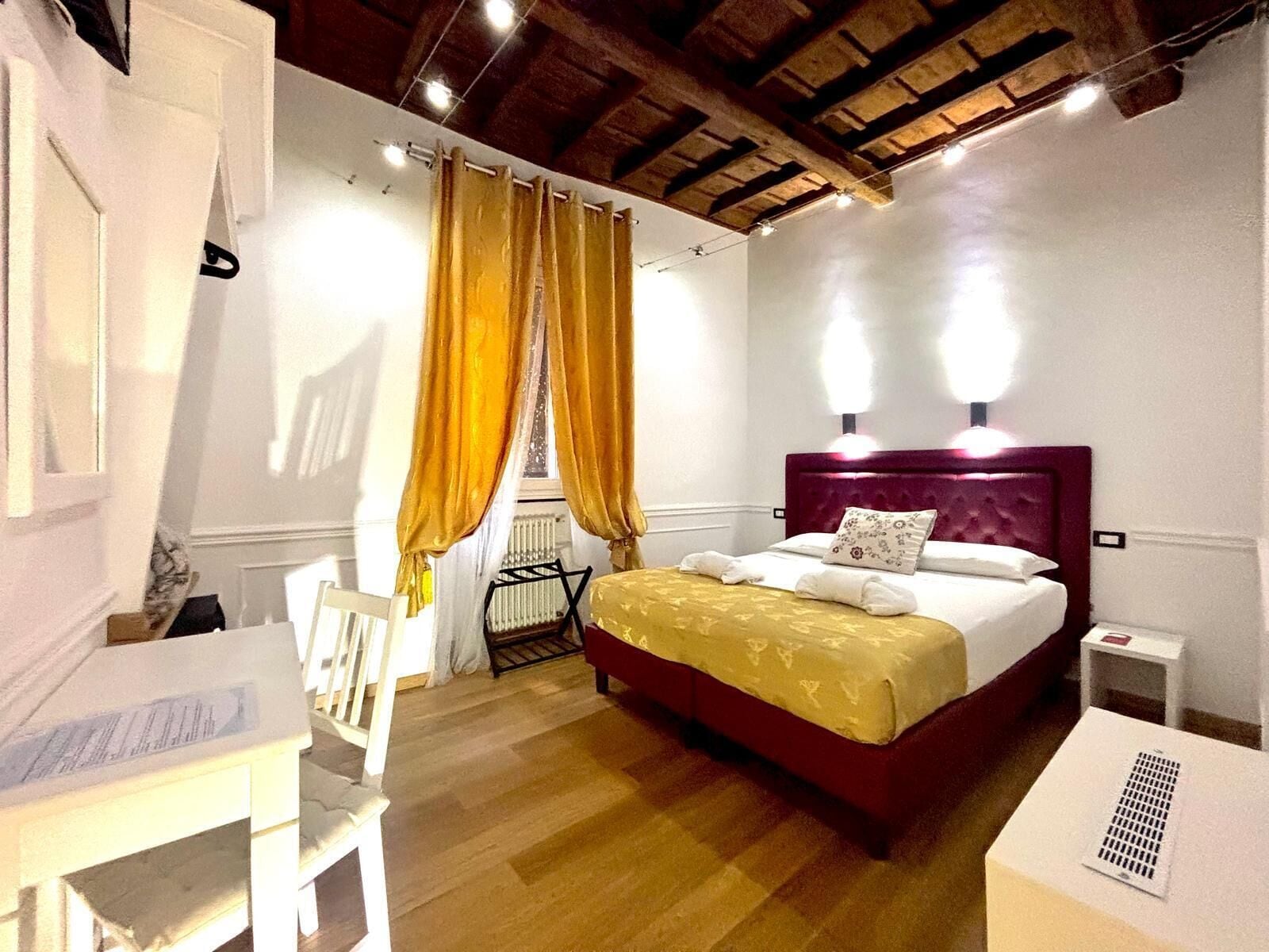 A hotel room with a wooden ceiling, a bed with a red headboard, a white bedside table, and a small white table.