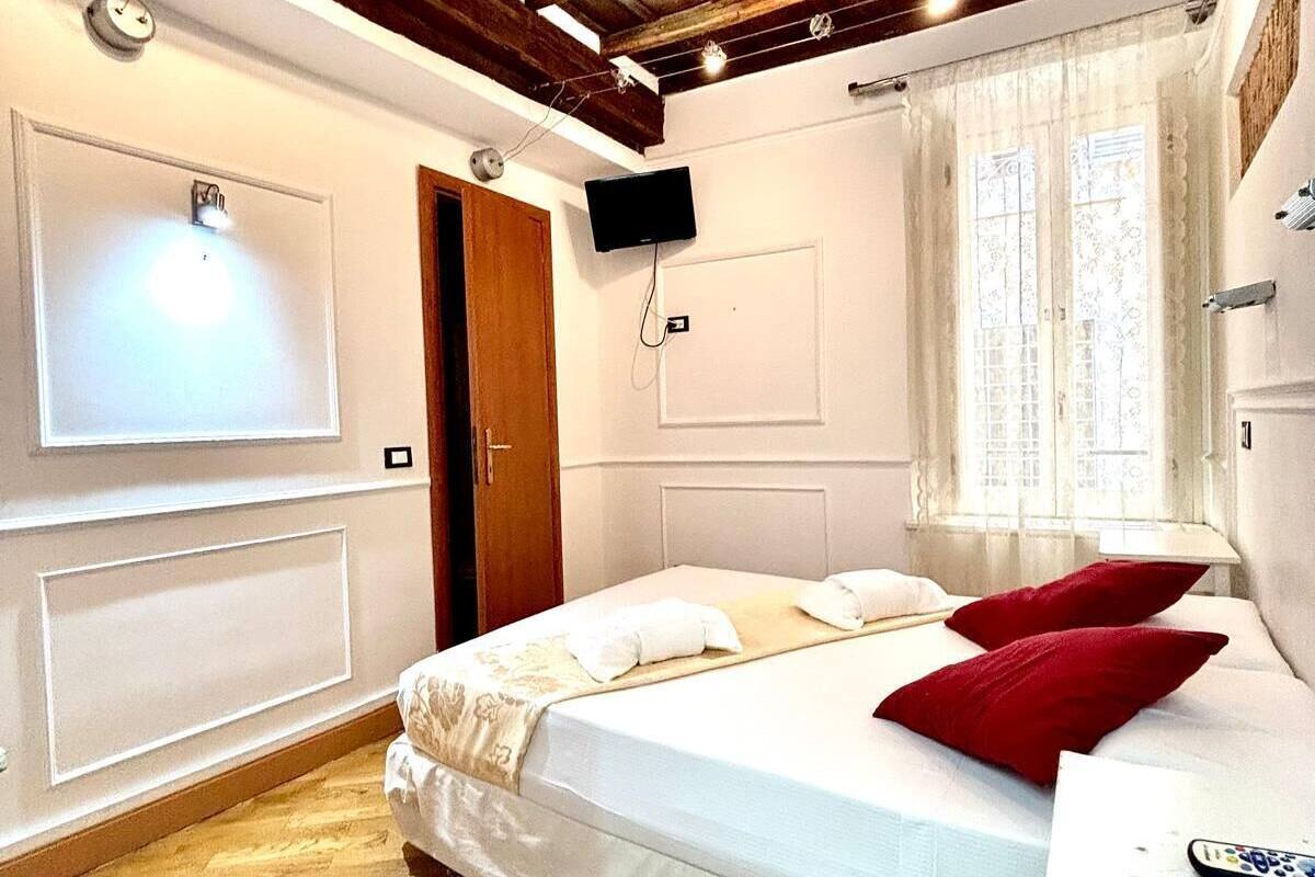 A bedroom with a wooden ceiling, a bed with white bedding and red pillows, a TV mounted on the wall, and a window with sheer curtains.
