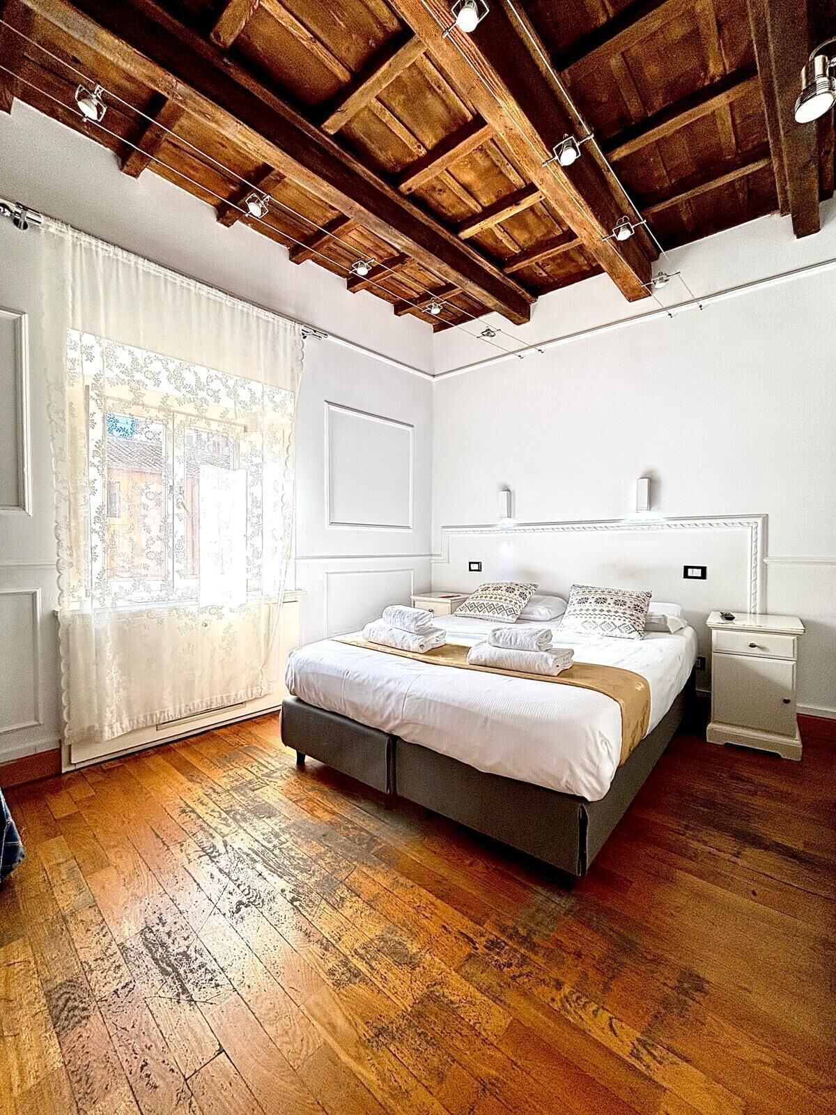 Ein Schlafzimmer mit Holzbalkendecke, einem Bett mit weißer Bettwäsche, einem Nachttisch und einem Fenster mit Vorhängen.