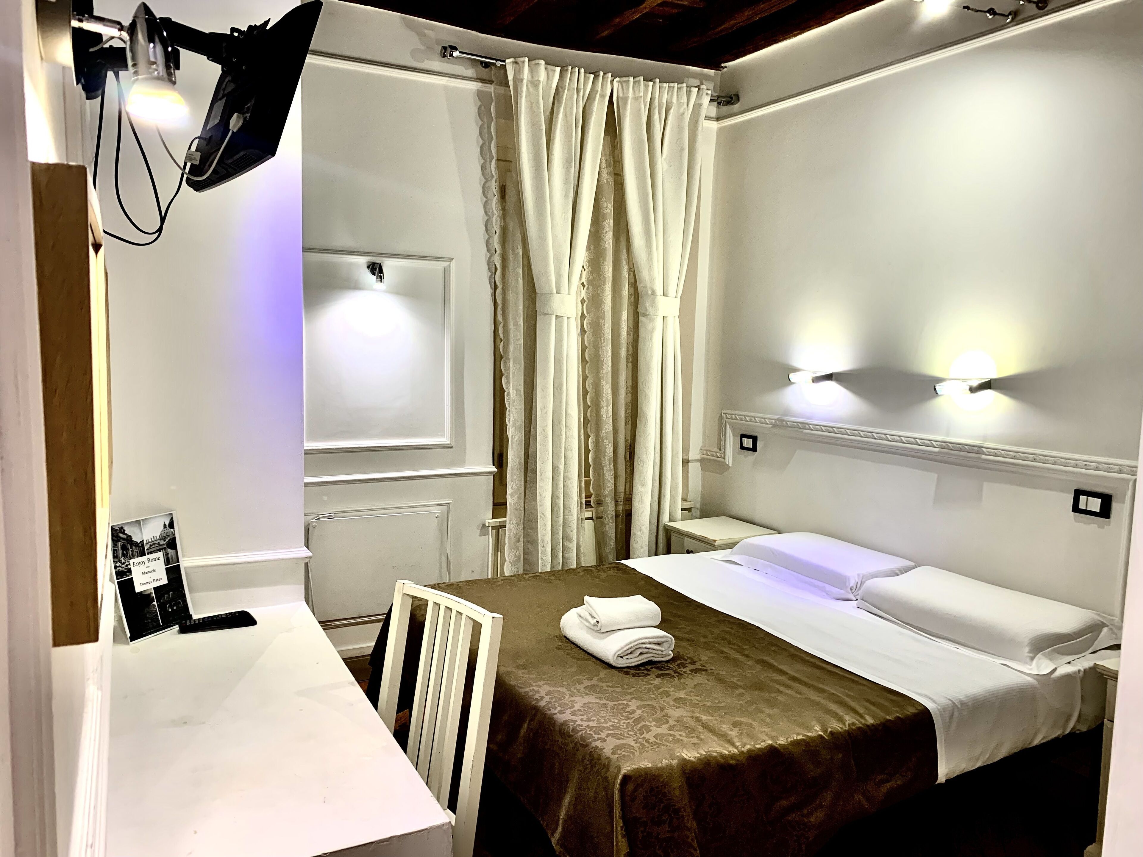 A hotel room with a bed, a nightstand, a desk with a lamp, and a window with curtains.