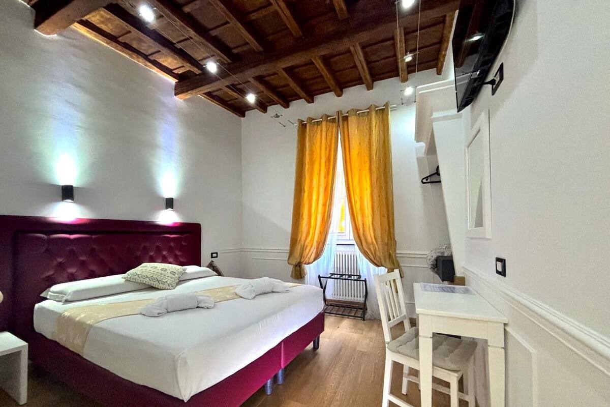 A bedroom with a red headboard, a bed with white linens, a desk with a chair, and a window with yellow curtains.