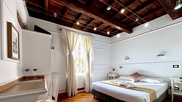 A bedroom with a wooden ceiling, a bed with white bedding, a bedside table with a lamp, and a window with curtains. - Domus Ester Roma (Rome)