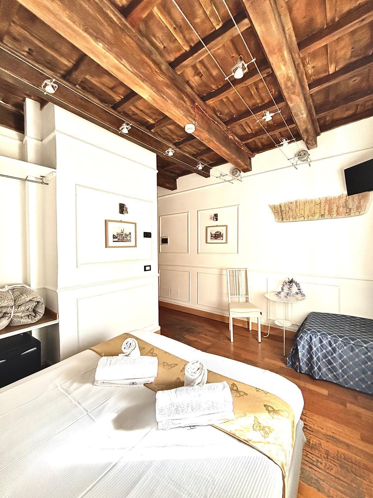 Una habitación con techo de madera, una cama con sábanas blancas, una silla, una mesita con un florero y cuadros en la pared.