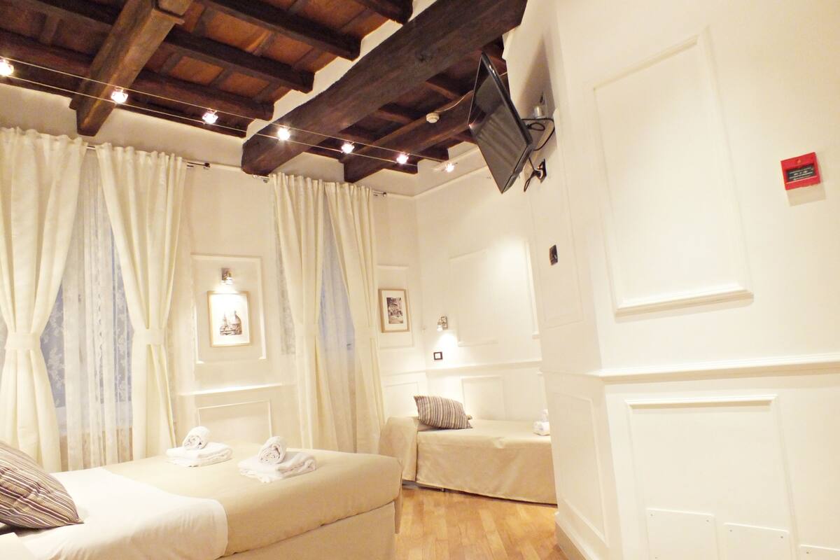 A hotel room with a bed, a smaller bed or sofa, wooden beams on the ceiling, and a flat-screen TV mounted on the wall.